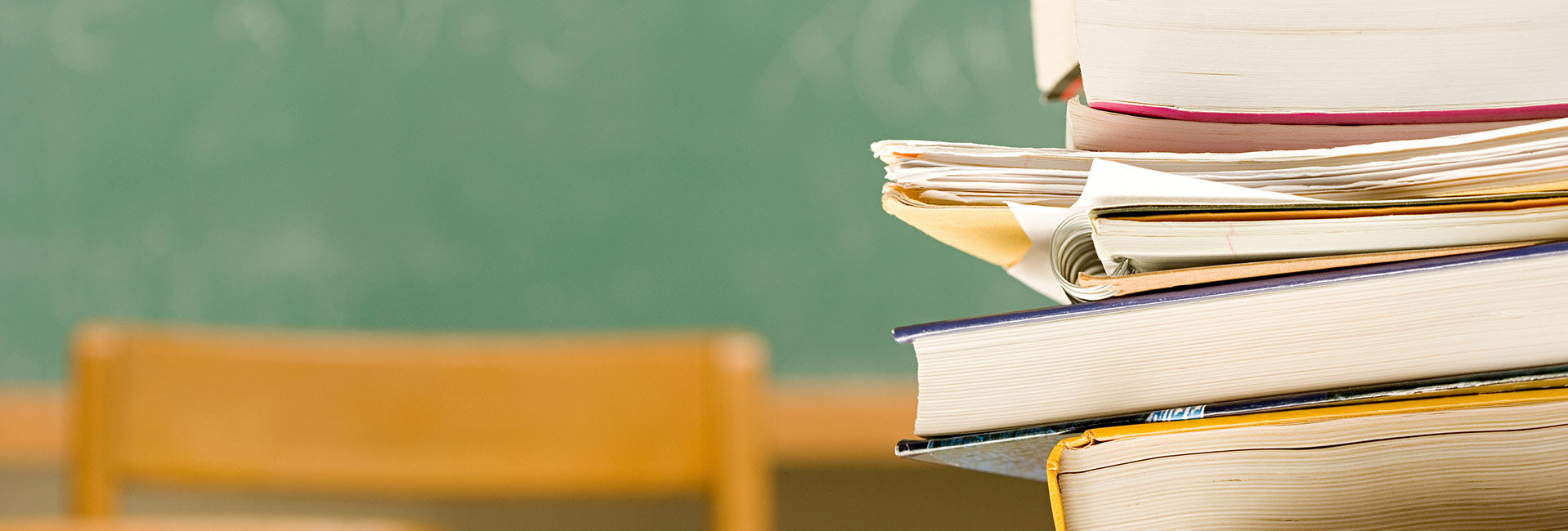 Das Bild zeigt auf der rechten Seiten einen Stapel Bücher, der im Vordergrund steht. Im Hintergrund ist eine Stuhllehne mit einer grünen Schultafel dahinter zu sehen.